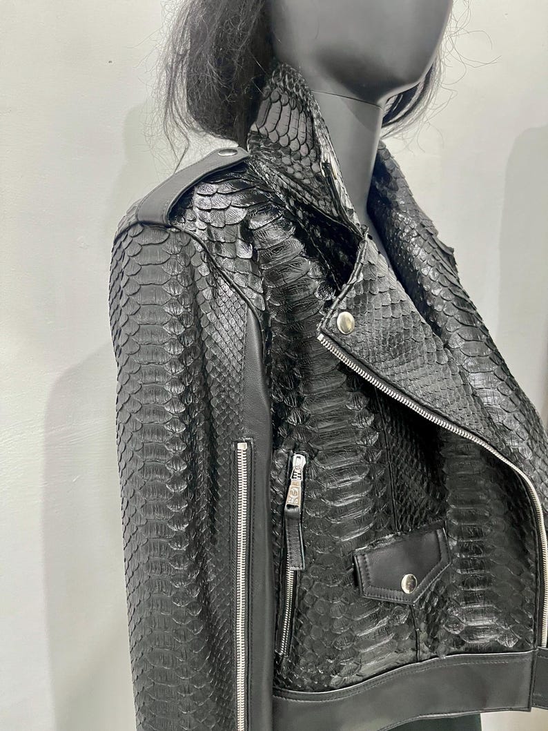 K&ouml;nnte beinhalten: Schwarze Lederjacke mit Schlangenhautmuster. Die Jacke hat einen Rei&szlig;verschluss und zwei Taschen.