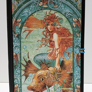 May include: A vintage-style book cover with a mermaid riding a fish with gears. The mermaid has long red hair and is wearing a green tail. The fish is orange and has large fins. The background is a teal blue with a floral pattern.