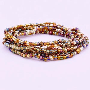 Könnte beinhalten: Ein mehrsträngiges Armband aus kleinen, runden Glasperlen in verschiedenen metallischen und irisierenden Ausführungen, darunter Bronze-, Kupfer-, Gold- und tiefe Violetttöne, angeordnet in einer kreisförmigen, gewickelten Form auf hellem Grund.