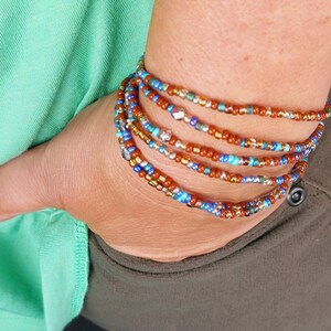 Könnte beinhalten: Ein Satz übereinander gelegter Perlenarmbänder in Braun-, Blau- und Goldtönen. Die Armbänder bestehen aus kleinen, runden Perlen und sind übereinander gestapelt.