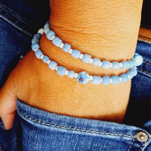 Könnte beinhalten: Zwei Armbänder mit hellblauen Perlen, weißen Akzenten und einem floralen Design. Die Armbänder werden an einem Handgelenk getragen, mit einem Jeans-Hintergrund.