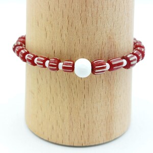 Könnte beinhalten: Ein rotes und weiß gestreiftes Perlenarmband mit einer einzelnen weißen Perle in der Mitte.