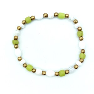 Op de afbeelding: Een cirkelvormige kralenarmband met een herhalend patroon van metallic gouden bolvormige kralen, matte limoengroene cilindrische kralen en gladde witte ovale kralen, gerangschikt in een doorlopende lus op een elastisch koord.