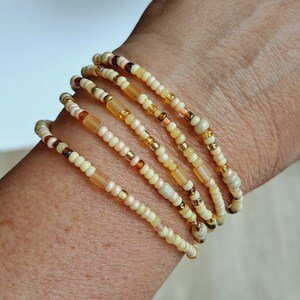 Könnte beinhalten: Ein mehrsträngiges Armband aus kleinen, runden Glasperlen in Creme-, Beige- und durchscheinenden Bernsteintönen. Das Design zeigt ein sich wiederholendes Muster aus undurchsichtigen und klaren Perlen, das einen geschichteten Look erzeugt.