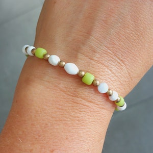 Op de afbeelding: Een armband gemaakt van witte en groene kralen met gouden accenten. De armband zit om een pols.