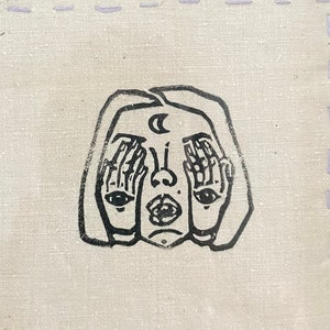 Op de afbeelding: Een ingelijst zwart-witte illustratie van een persoon die zijn ogen bedekt met zijn handen. De illustratie is op een beige stoffen achtergrond met een lichtpaarse gestikte rand.