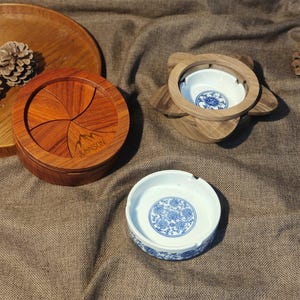Op de afbeelding: Een ronde houten doos met een draaibaar deksel met een bergafbeelding en de tekst JOHNSON, naast een houten houder met een witte keramische kom met blauwe bloemmotieven en een losse witte keramische kom met blauwe bloemmotieven.