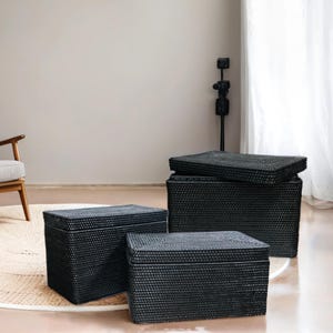 May include: Three black woven storage baskets of varying sizes. The baskets are made of a natural material and have a textured surface. The baskets are arranged in a staggered formation on a light brown rug.