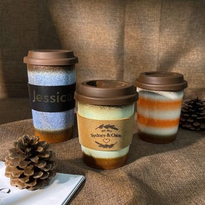 May include: Three ceramic travel mugs with brown lids. The mugs have different designs. One mug has a blue and brown design with the name "Jessica" on it. Another mug has a brown, white, and orange design with the text "Est. 2019 Sydney & Chloe" on it. The third mug has a brown, white, and orange design.