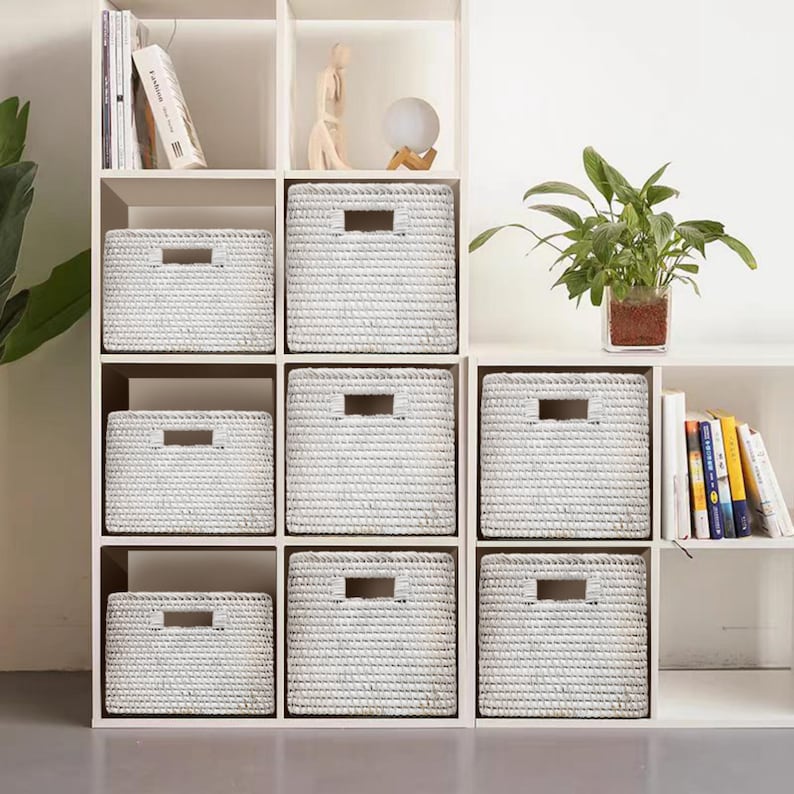 May include: White wicker storage baskets on a white shelf unit. The baskets are all the same size and have a woven pattern. The shelf unit has three shelves and is made of wood.