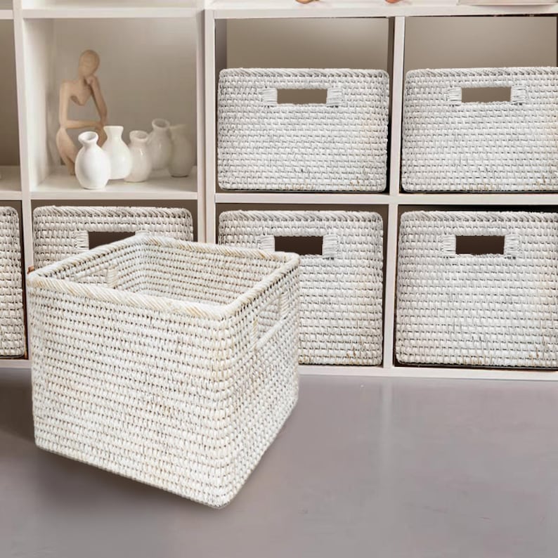 May include: White wicker storage baskets with handles, arranged on a white shelf unit. The baskets are woven with a fine, intricate pattern.