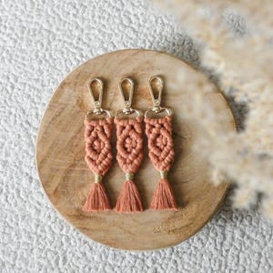 Peut inclure: Trois porte-clés macramé pêche, chacun avec un fermoir doré et une pampille assortie. Ils sont présentés sur une assiette en bois ronde, mettant en valeur un style bohème. Les porte-clés mesurent environ 13 cm de long.