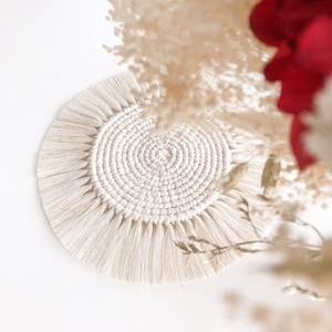 Peut inclure: Un dessous de verre rond en macramé blanc cassé, avec un motif en spirale au centre et des franges sur les bords. Le dessous de verre est en coton tissé et est présenté sur un fond blanc. Des fleurs séchées blanches et rouges sont en arrière-plan.