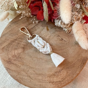 Puede incluir: Un llavero de macramé hecho a mano con un cierre dorado y un pequeño acento de concha. El diseño tejido en blanco roto presenta una borla dorada y un pequeño acento dorado. El llavero se muestra sobre una superficie de madera con flores secas en el fondo.