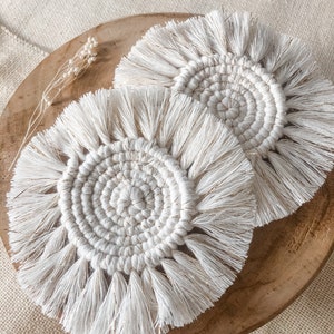 Peut inclure: Deux dessous de verre en macramé blanc avec un motif circulaire et des franges. Les dessous de verre sont posés sur une assiette en bois, avec un tissu de couleur neutre en arrière-plan. Les dessous de verre ont un centre en spirale et des accents dorés.