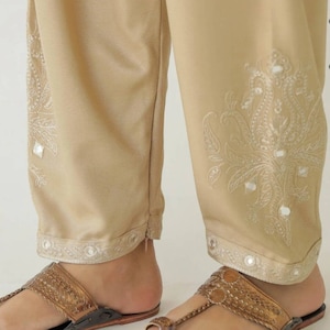 May include: Beige pants with white embroidery and mirror embellishments. The pants have a wide leg and a fitted waist. The pants are worn with brown leather sandals.