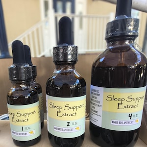 May include: Several amber glass bottles of Sleep Support Extract are displayed. The bottles have black droppers and labels with the text "Sleep Support Extract" and the size in fluid ounces: 1 fl oz, 2 fl oz, and 4 fl oz. The brand name "Amber Rose Apothecary" is also visible.