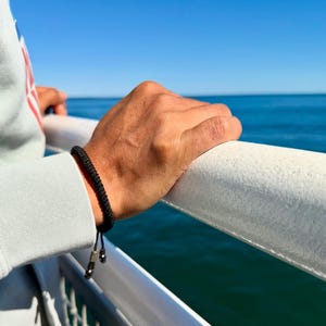 May include: A black braided bracelet with silver-colored beads is worn on a wrist. The person is holding onto a white railing, with a view of the ocean and blue sky in the background.