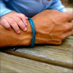 May include: A close-up of a person's wrist wearing a teal blue braided bracelet. The bracelet is made of multiple strands of cord and has a small black bead at the end.