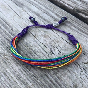 May include: A rainbow colored friendship bracelet with a purple cord and black beads. The bracelet has a tag that says "RUMI SUMAQ".
