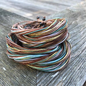 Peut inclure: Gros plan sur une pile de bracelets color&eacute;s en cordons fins avec des accents dor&eacute;s. Les bracelets sont dispos&eacute;s en cercle et sont faits de cordons bruns, blancs et turquoise.