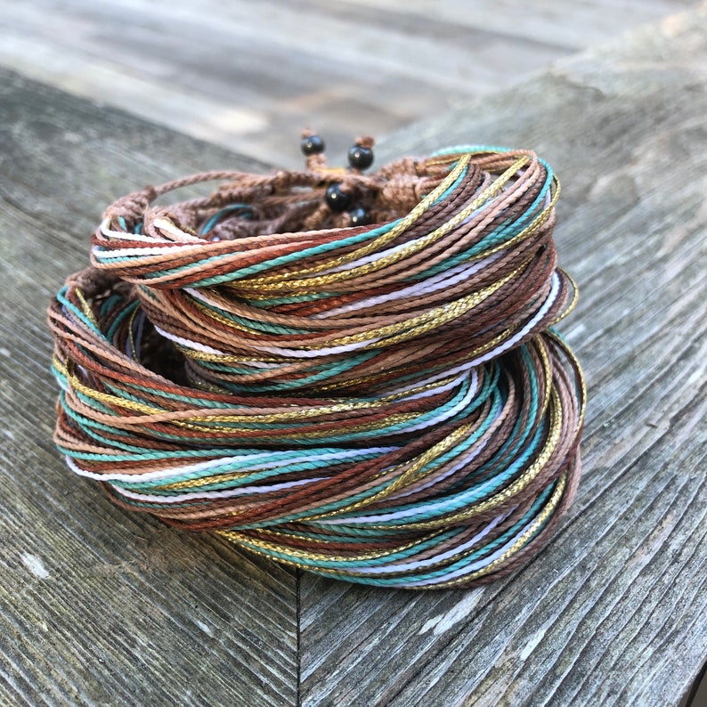 Peut inclure: Gros plan sur une pile de bracelets color&eacute;s en cordons fins avec des accents dor&eacute;s. Les bracelets sont dispos&eacute;s en cercle et sont faits de cordons bruns, blancs et turquoise.