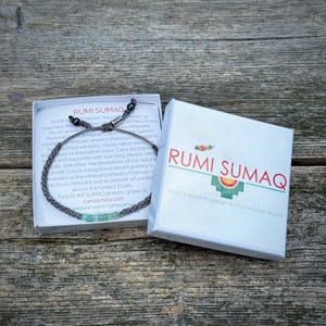 Custom handmade opal rope bracelet by Rumi Sumaq displayed in a branded gift box, perfect for men, women, and kids. Features blue green Peruvian Opal stones along hand-knotted fiber cords in personalized colors, finished with Hematite stone accents and a Rumi Sumaq logo bead. Ideal as a birthday, holiday, friendship, or meaningful gift, handcrafted by indigenous Quechua designer Coco Paniora Salinas on Martha’s Vineyard, combining artisan Peruvian heritage with wearable fiber art jewelry.