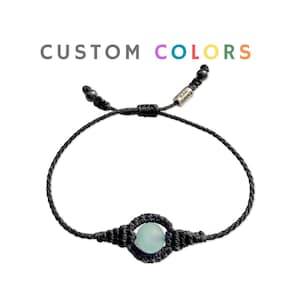 Puede incluir: Una pulsera de macramé negra con una piedra redonda azul claro en el centro. La pulsera tiene un cierre de nudo corredizo. El texto "CUSTOM COLORS" está encima de la pulsera en gris, morado, verde azulado y naranja.