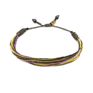 Puede incluir: Un brazalete trenzado multicolor con hilos amarillos, morados y marrones. El brazalete tiene un cierre de nudo deslizante.