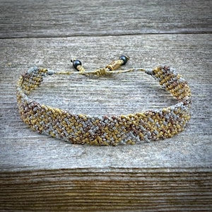 Op de afbeelding: Een geweven armband met een mix van gouden, bruine en grijze draden. De armband heeft een getextureerde uitstraling en is verstelbaar met een schuifknoopsluiting. De armband is ongeveer 1,3 cm breed.