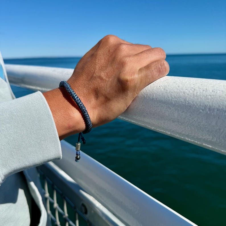 A Rumi Sumaq men’s blue rope bracelet is pictured on the Martha’s Vineyard ferry, a thoughtful handmade gift for him inspired by the sea. The adjustable waxed cord bracelet features Hematite stones and a Rumi Sumaq logo bead, visible as the man’s wrist rests on the ferry railing with the ocean beyond. A perfect men’s jewelry gift that blends coastal charm, fine craftsmanship, and the artistry of indigenous Quechua Peruvian designer Coco Paniora Salinas.
