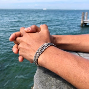 May include: A close-up of a person's wrist wearing a multi-colored braided bracelet. The bracelet is made of a combination of white, gray, and black threads. The person's hand is resting on a wooden surface.