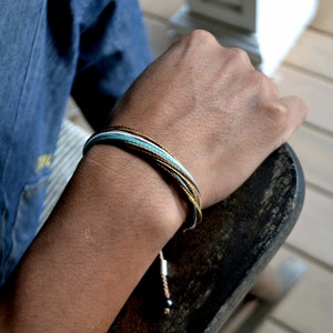 Peut inclure: Un bracelet en cordelette multicolore avec des fils marrons, blancs et bleus. Le bracelet est nou&eacute; et comporte une petite perle noire &agrave; l'extr&eacute;mit&eacute;.