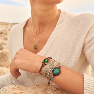 Peut inclure: Une femme portant un pull blanc avec un collier en or et un pendentif en pierre verte. Elle porte plusieurs bracelets au poignet, dont un avec une grosse pierre verte.