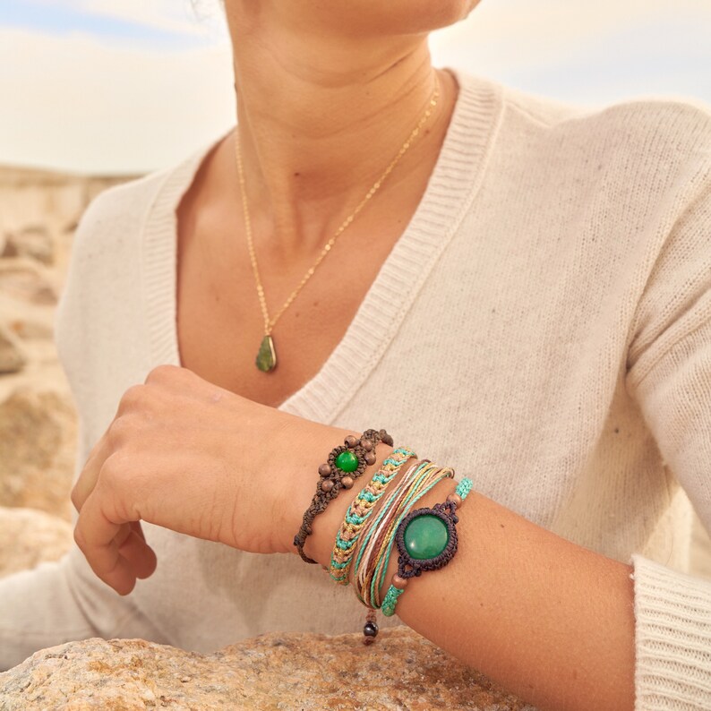 Peut inclure: Une femme portant un pull blanc avec un collier en or et un pendentif en pierre verte. Elle porte plusieurs bracelets au poignet, dont un avec une grosse pierre verte.
