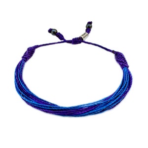 Op de afbeelding: Een blauw en paars geweven armband met een verstelbare schuifknoopsluiting. De armband is gemaakt van meerdere strengen blauwe en paarse draden. De merknaam "SUMAQ" is zichtbaar op een klein metalen label.