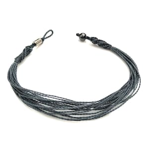 Puede incluir: Un brazalete de cordón trenzado gris con una cuenta negra y un cierre plateado.
