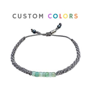 May include: A gray braided bracelet with a row of light green beads. The bracelet has a sliding knot closure and small black beads at each end. The text "CUSTOM COLORS" is above the bracelet in gray, purple, teal, yellow, and orange.