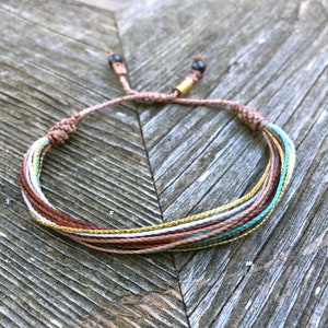 Peut inclure: Un bracelet tress&eacute; multicolore avec des fils bruns, blancs, dor&eacute;s et turquoise. Le bracelet est dot&eacute; de perles noires aux extr&eacute;mit&eacute;s et d'un accent en m&eacute;tal dor&eacute;.