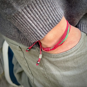 May include: A red, green, and red braided bracelet with a small silver charm and black beads. The bracelet is worn on a wrist, with a gray sweater and green pants in the background.