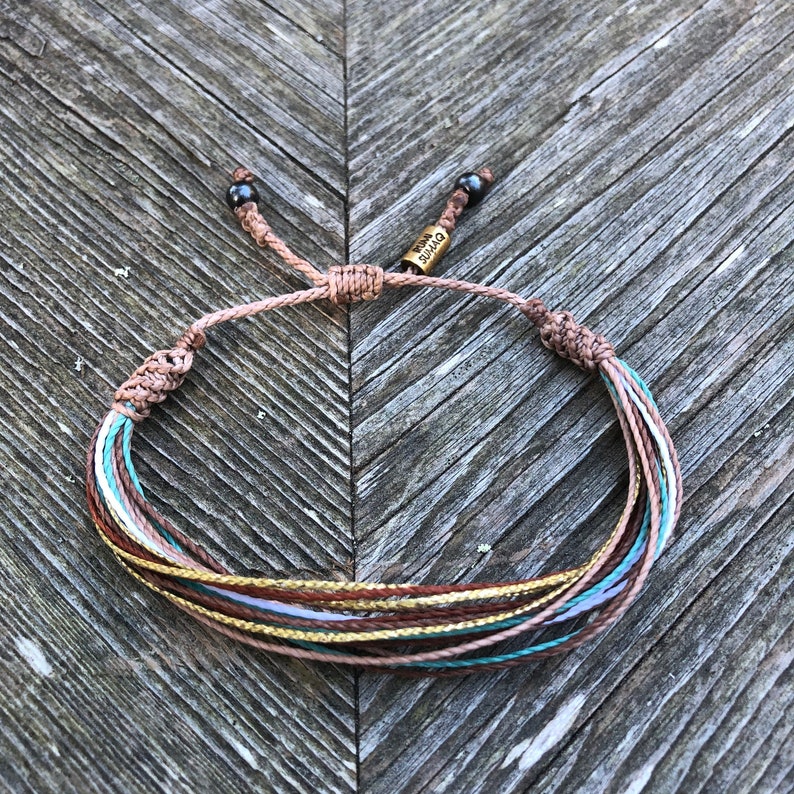 Peut inclure: Un bracelet tress&eacute; multicolore avec un cordon marron et une &eacute;tiquette m&eacute;tallique sur laquelle est inscrit "Divine". Le bracelet est nou&eacute; avec deux perles noires.