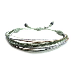 May include: A multi-strand bracelet with green, brown, white, and silver threads. The bracelet is tied with a knot and has two small black beads at the ends.