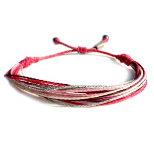 Puede incluir: Pulsera de la amistad multicolor con hilos rosas, rojos, blancos y plateados. La pulsera está atada con un nudo en cada extremo.