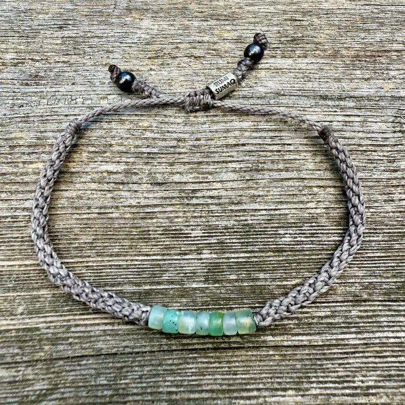 Flat lay of a custom handmade rope bracelet by Rumi Sumaq photographed on a weathered coastal deck board, showcasing the earthy texture of hand-knotted fiber cords chosen in personalized colors. Centered blue green Peruvian Opal stones evoke ocean tones, while Hematite beads accent the adjustable drawstring tails beside the Rumi Sumaq logo bead. Handcrafted by Quechua designer Coco Paniora Salinas on Martha’s Vineyard, this unisex bracelet captures coastal elegance and artisan heritage.