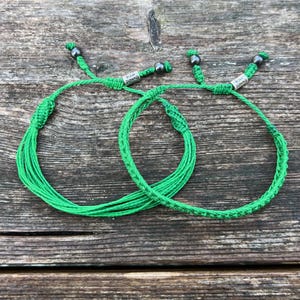 May include: Two green friendship bracelets with silver beads and the text "RUMI SUMAQ" on each bracelet.