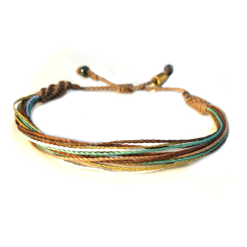 Peut inclure: Un bracelet multi-brins avec des fils de couleur marron, blanc, or et vert. Le bracelet est nou&eacute; &agrave; l'extr&eacute;mit&eacute;.