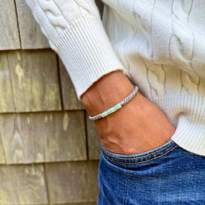 Custom color handmade rope bracelet by Rumi Sumaq shown on a man’s wrist styled with dark jeans and a white sweater, highlighting the casual sophistication of artisan jewelry. The coastal bracelet features a line of genuine blue green Peruvian Opal stones framed by hand-knotted fiber cords in the customer’s chosen colors. Hematite stone accents and a Rumi Sumaq logo bead complete the adjustable design handcrafted by indigenous Quechua jewelry designer Coco Paniora Salinas on Martha’s Vineyard.