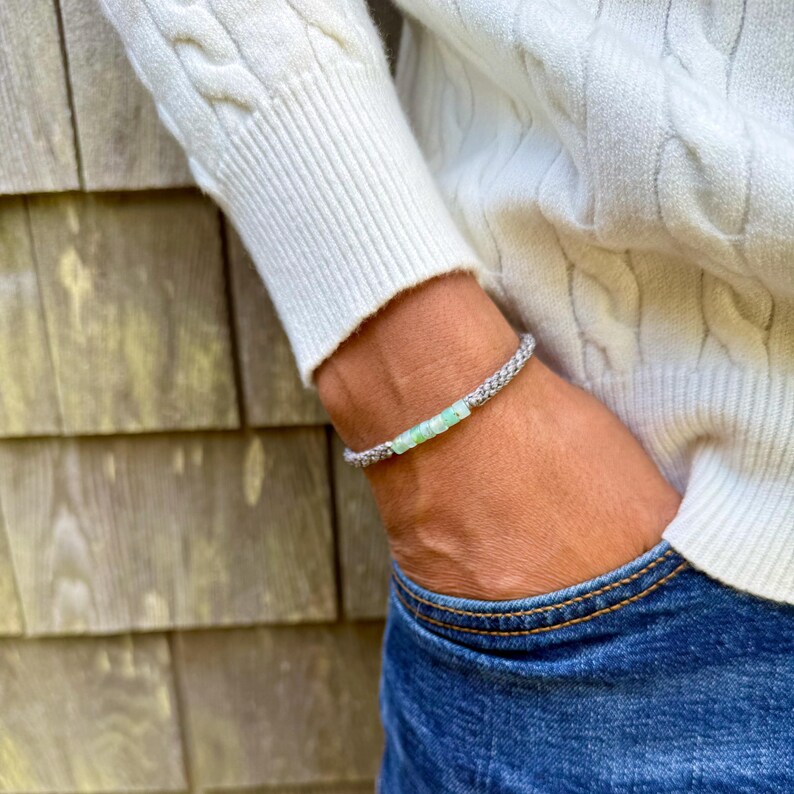 Custom color handmade rope bracelet by Rumi Sumaq shown on a man’s wrist styled with dark jeans and a white sweater, highlighting the casual sophistication of artisan jewelry. The coastal bracelet features a line of genuine blue green Peruvian Opal stones framed by hand-knotted fiber cords in the customer’s chosen colors. Hematite stone accents and a Rumi Sumaq logo bead complete the adjustable design handcrafted by indigenous Quechua jewelry designer Coco Paniora Salinas on Martha’s Vineyard.