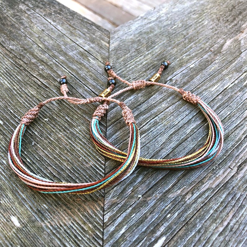 Peut inclure: Deux bracelets d'amiti&eacute; multicolores avec des fils marrons, blancs, dor&eacute;s et turquoise. Les bracelets sont nou&eacute;s avec des perles noires et des fermoirs en m&eacute;tal dor&eacute;.