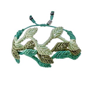 May include: Green and light green macrame bracelet with a leaf design. The bracelet has black beads on the ends and a silver tag that says "OMNES".